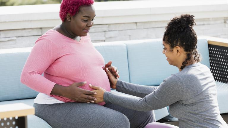 Pregnant person in pink blouse and red hair sitting on a blue couch with a doula holding their belly. Both are smiling.