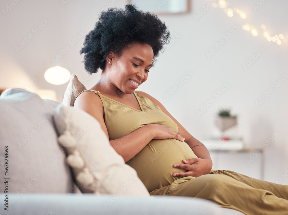 Black pregnant woman sitting on a white couch. She is smiling.